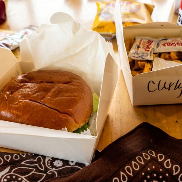 A burger and curly fries in takeout containers, with chips and a Pepsi on a wooden table.