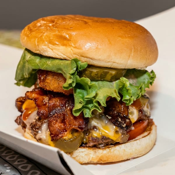 A gourmet burger with bacon, cheese, lettuce, pickles, and onion rings in a white paper tray.