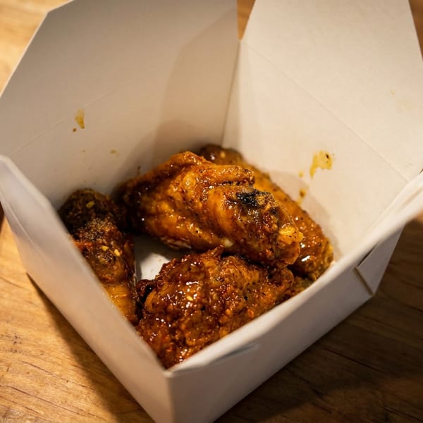 Crispy chicken wings in a white takeout box next to a bag of chips on a wooden table.